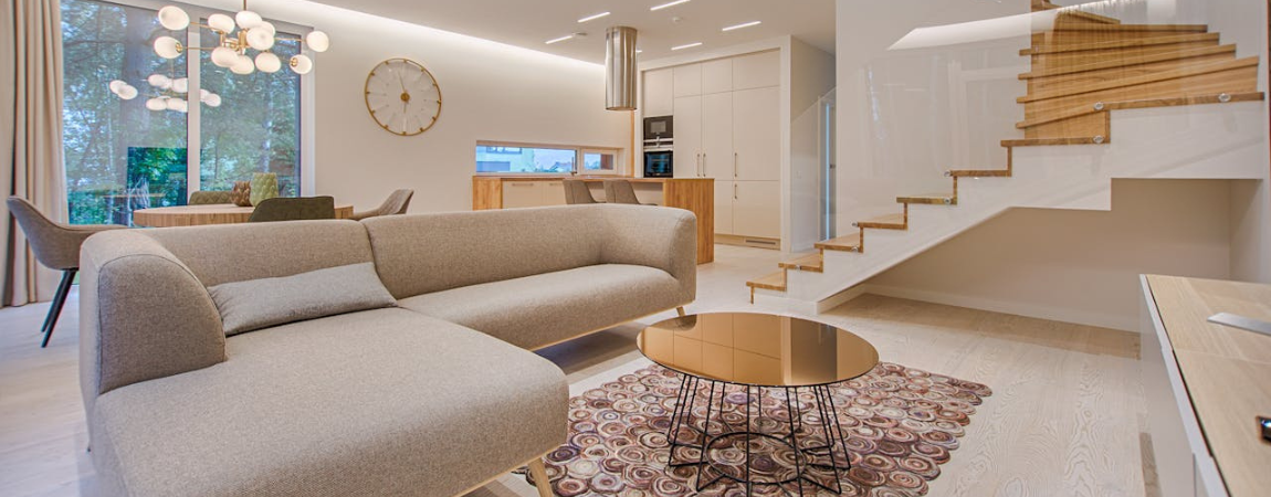 Modern minimalist living room with a beige sofa, round metallic coffee table, patterned rug, wooden staircase with glass railing, and open kitchen area.